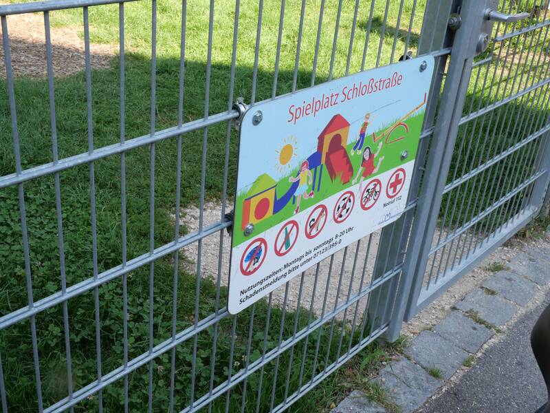An einem Metalltor hängt ein Schild mit der Aufschrift „Spielplatz Schloßstraße“ und Piktogrammen mit Regeln.