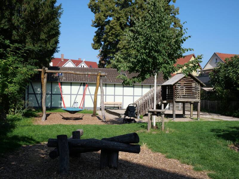 Ein Holzhaus mit Dach und Rutsche steht auf einem Sandbereich, daneben eine Nestschaukel vor Wohnhäusern.