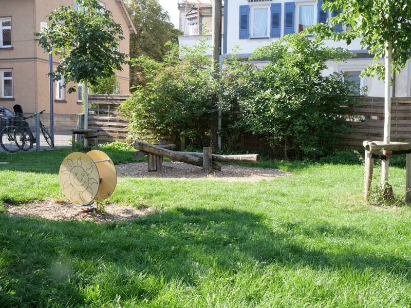 Eine kleine Wippe aus Holzstämmen steht im Gras, im Vordergrund eine gelbe Spieltrommel mit Sonnengesicht.