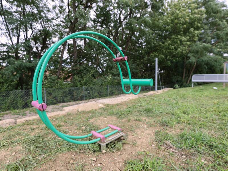 Ein grünes Metallspielgerät mit Haltegriffen und einer kleinen Sitzfläche steht auf einer Grasfläche, im Hintergrund ein Zaun und Bäume.