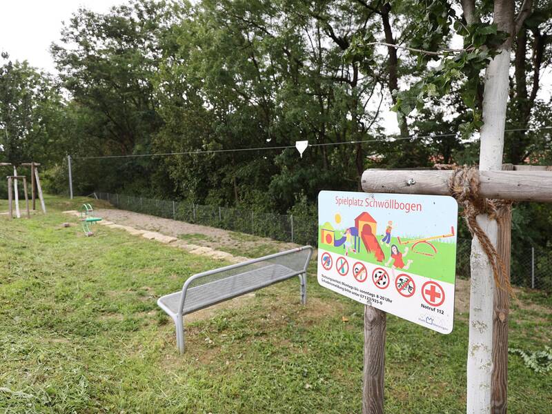 Ein Spielplatzschild mit der Aufschrift „Spielplatz Schwöllbogen“ steht neben einer Bank, im Hintergrund eine Seilbahn, ein Klettergerät und eine Rasenfläche.