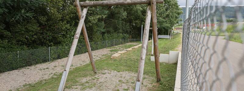 Eine große Holz-Konstruktion für eine Seilbahn steht auf einer leicht abschüssigen Wiese, daneben ein Kiesweg und ein Zaun.