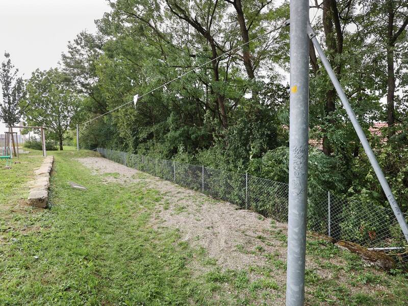 Eine Seilbahn mit langem Kiesweg führt bergab, daneben eine Wiese mit Steinen, Bäumen und einem Zaun.