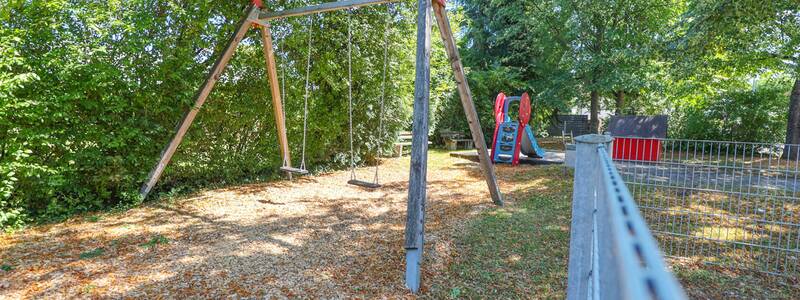 Eine Schaukel mit Holzgestell und zwei Schaukelsitzen steht auf einem mit Holzschnitzeln bedeckten Spielbereich, im Hintergrund die Rutsche und ein rotes Spielhaus.