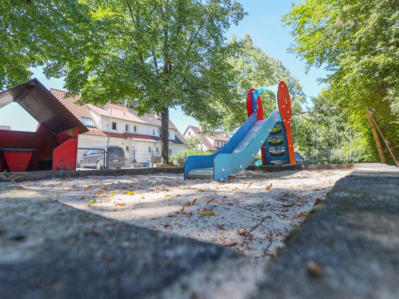 Eine blaue Rutsche mit roten Seitenteilen steht auf einem Sandspielbereich, daneben ein kleines rotes Spielhaus mit Dach, im Hintergrund Bäume und Wohnhäuser.