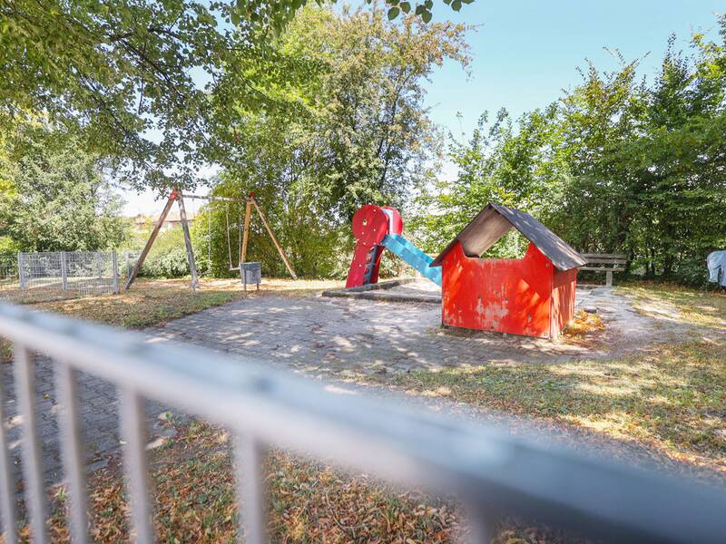 Ein eingezäunter Spielplatz mit einer Rutsche, einer Schaukel und einem roten Spielhaus, umgeben von Bäumen und Büschen.