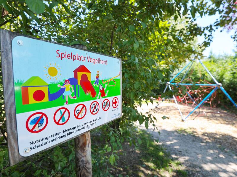 Ein Spielplatzschild mit der Aufschrift „Spielplatz Vogelherd“ steht vor einer Kletterpyramide aus Seilen, umgeben von Bäumen und Sträuchern.