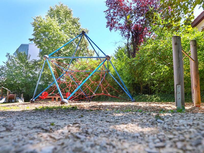 Eine große Kletterpyramide aus blauen Metallstangen und roten Seilen steht auf einem mit Holzschnitzeln bedeckten Spielbereich, umgeben von Bäumen.