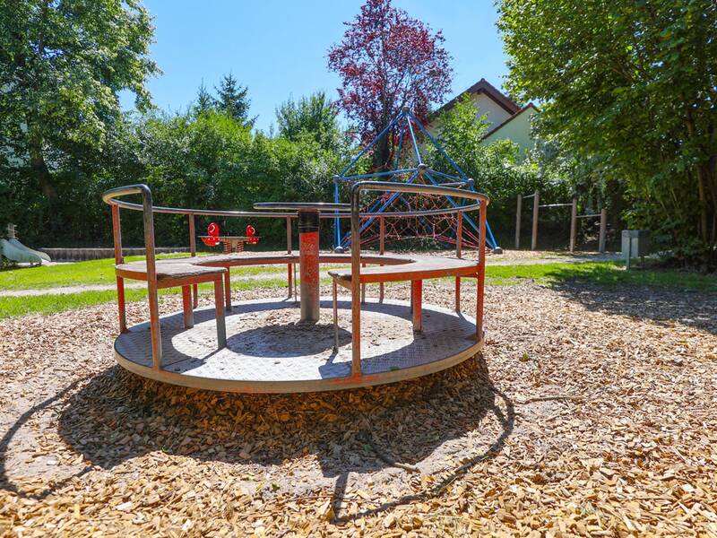 Ein Karussell mit Metallgestell und Holzsitzen steht auf einem Spielplatz mit Holzschnitzelboden, im Hintergrund eine Kletterpyramide und weitere Spielgeräte.