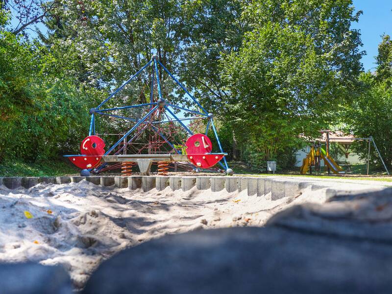  Ein Sandkasten mit Begrenzungssteinen liegt vor einer Kletterpyramide und einer Wippe mit roten Sitzen, umgeben von Bäumen und Sträuchern.