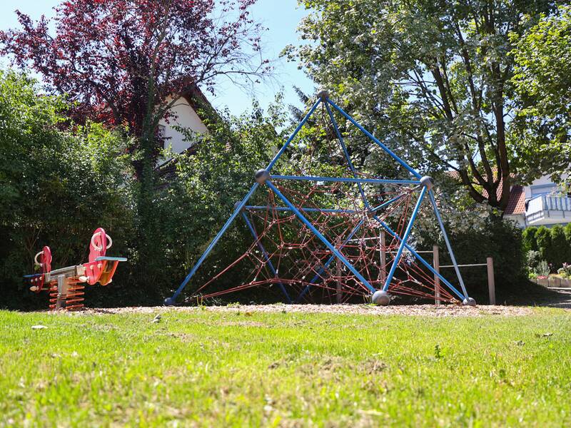 Eine Kletterpyramide aus Metallstangen und Seilen steht auf einem mit Holzschnitzeln bedeckten Spielplatz, daneben eine rote Federwippe, im Hintergrund Wohnhäuser und Gärten.