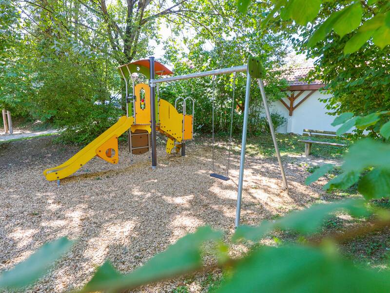 Ein gelbes Klettergerüst mit einer Rutsche und einer Schaukel steht auf einem schattigen Spielplatz mit Holzschnitzelboden, daneben eine Sitzbank.