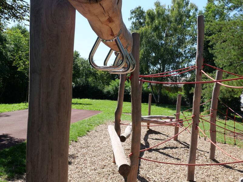 Zu sehen ist ein Holz-Klettergerüst mit Seilen in einem sonnigen Spielplatzbereich, umgeben von Bäumen und Gras.