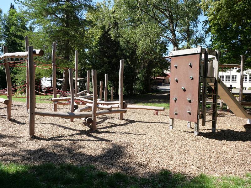 Ein Spielplatz aus Holz mit Klettergerüst, Kletterwand und Rutsche, umgeben von Bäumen.