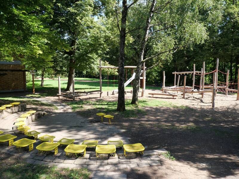 Ein Spielplatz im Grünen mit Klettergerüsten, Schaukeln und gelben Sitzbänken im Vordergrund.
