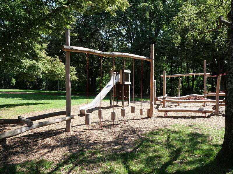 Ein Spielplatz mit Holzklettermöglichkeiten, Rutsche und Balancierstrecke befindet sich auf einer Rasenfläche, umgeben von Bäumen.