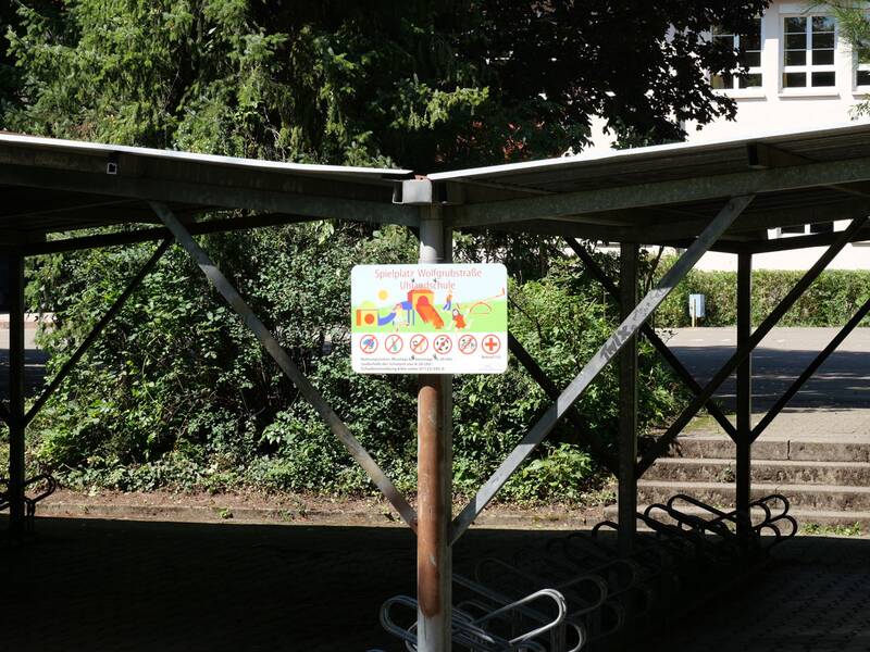 Ein Schild mit der Aufschrift "Spielplatz" und einigen Piktogrammen hängt unter einem überdachten Fahrradständer, umgeben von Bäumen und einem Gebäude im Hintergrund.
