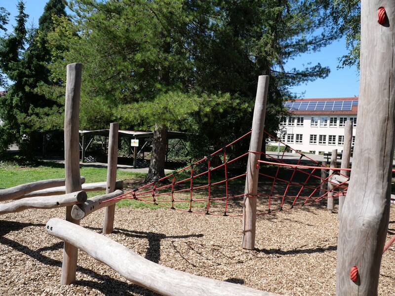 Ein Klettergerüst mit Netz aus Seilen und Holzbalken steht auf einem mit Holzspänen bedeckten Spielplatz, umgeben von Bäumen, mit einem Gebäude im Hintergrund.