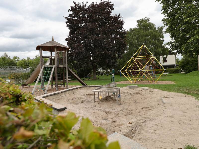Ein Spielplatz mit einer Rutsche, einem Klettergerüst und einem großen Sandkasten, umgeben von Bäumen und Rasenflächen.