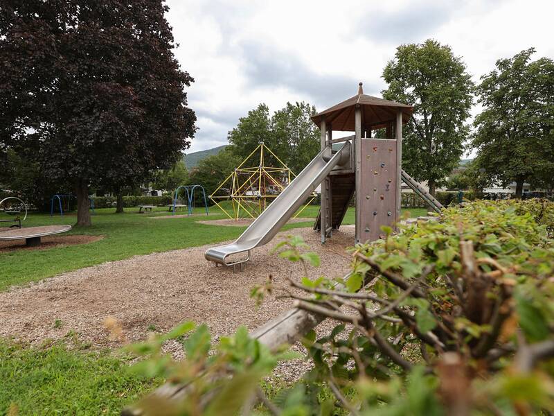 Ein großer Spielturm mit Rutsche, Kletterwand und Leiter steht auf einem Spielplatz, im Hintergrund eine gelbe Kletterpyramide und Schaukeln.
