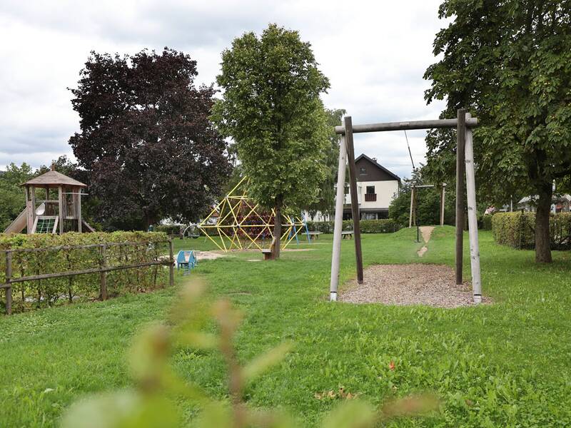 Blick auf eine Seilrutsche, Kletterpyramide, Schaukeln und Spielturm, eingebettet in eine grüne Wiese mit Bäumen.