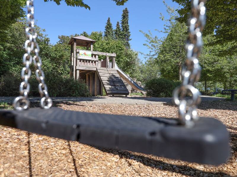 Der Blick durch eine Schaukel zeigt ein Holzspielhaus mit Kletterwand und Rutsche in einem grünen, offenen Bereich.