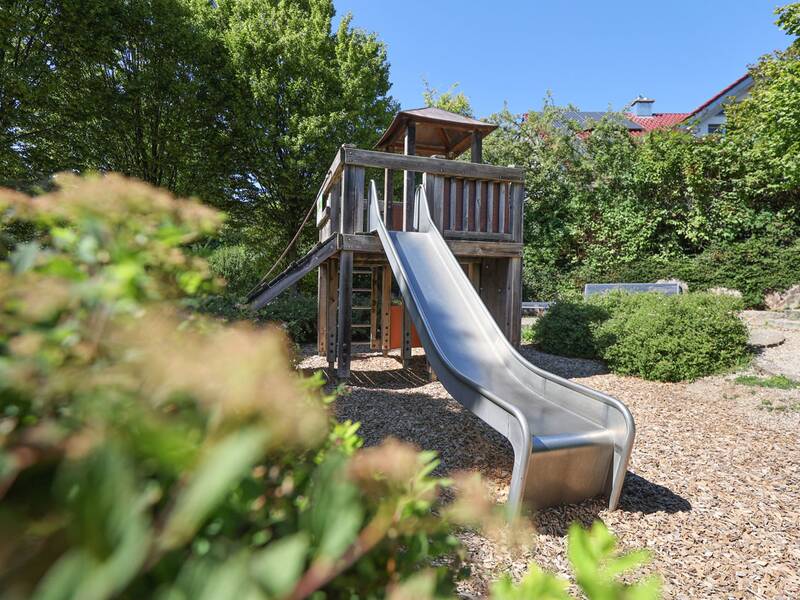 Eine Rutsche aus Metall ragt von einem Holzspielhaus hinunter, umgeben von Büschen und Mulchboden.