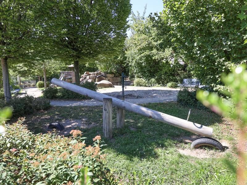 Eine Holzwippe steht in einem schattigen, bewachsenen Bereich mit Blick auf ein Spielgerüst und eine Felsenlandschaft.