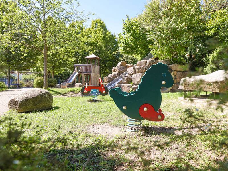 Im Vordergrund steht ein Wipptier in Pferdeform, im Hintergrund sind ein Holzklettergerüst mit Rutsche und eine große Steinformation mit zweiter Rutsche zu sehen.