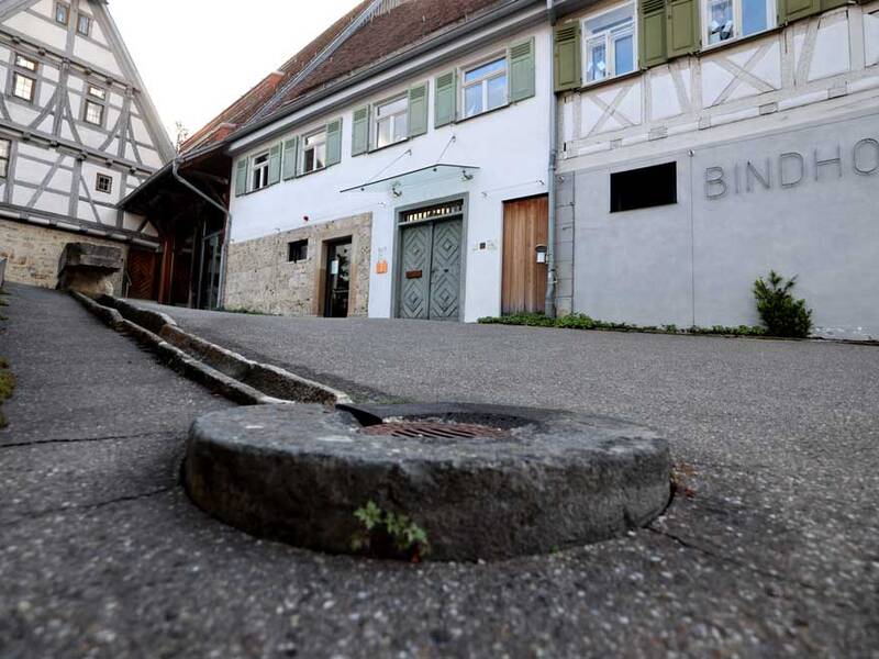 Ein traditionelles Fachwerkhaus steht neben einem Gebäude mit der Aufschrift "BINDHOF", mit einem kleinen Wasserkanal auf dem gepflasterten Weg davor.