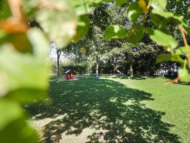 Freibad Metzingen, Blick durch ein Gebüsch auf den Spielbereich, Wiese liegt teilweise im Schatten