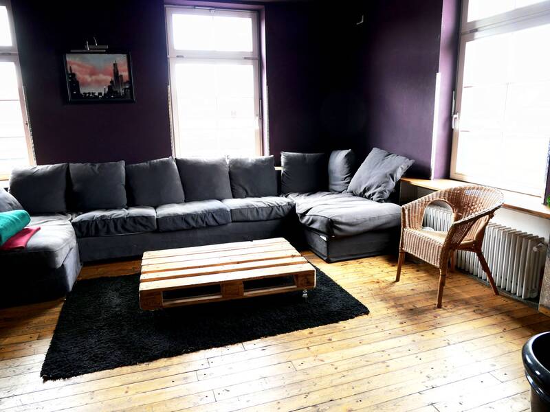 Jugendhaus. Ein gemütliches Zimmer mit einer großen, grauen Ecksofa, einem niedrigen Tisch aus Paletten, einem schwarzen Teppich und einem Korbstuhl, umgeben von dunklen Wänden und hellen Fenstern.