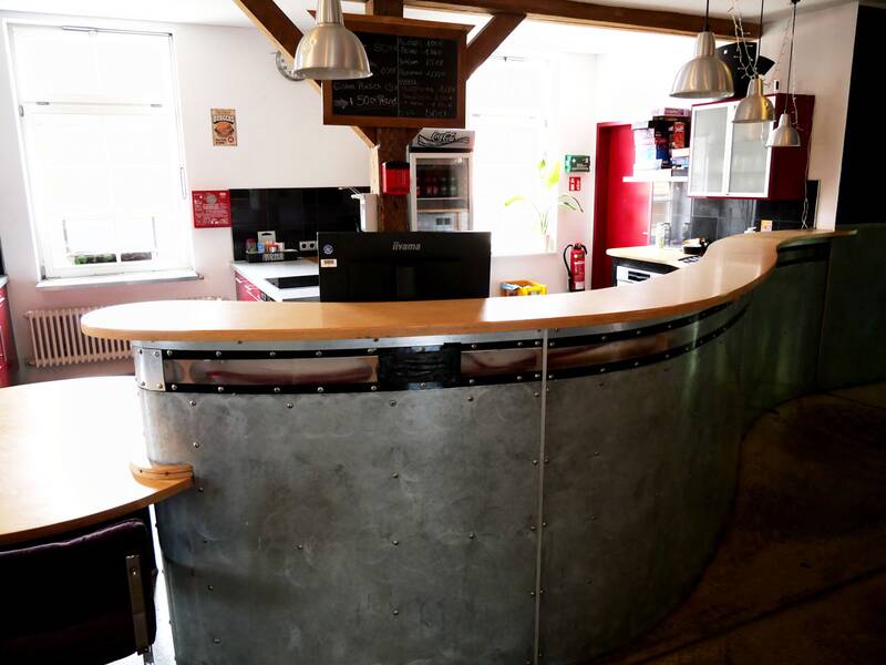 Jugendhaus. Ein halbrunder Tresen aus Holz und Metall steht in einem modern ausgestatteten Raum mit Küchenzeile und mehreren Hängelampen.