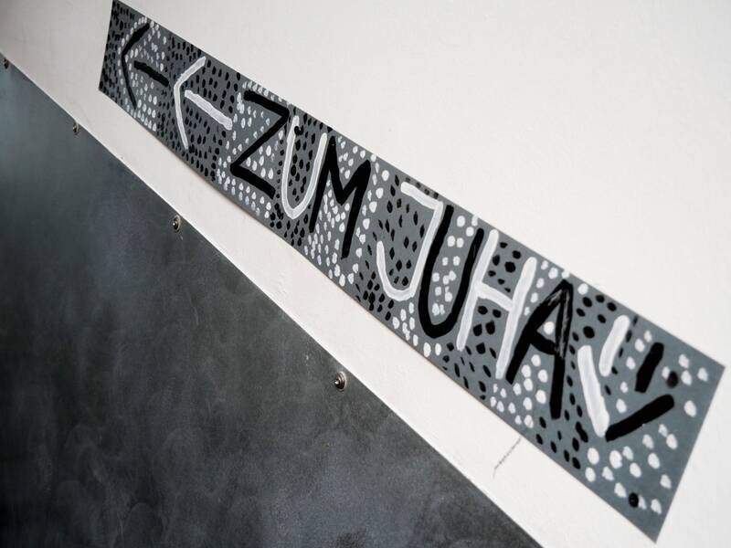 Jugendhaus. Ein handgemaltes Pfeilschild weist den Weg "Zum Juha" auf einem grauen, punktverzierten Hintergrund.