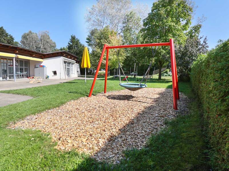 Ein Spielplatz mit einer roten Schaukel in Nestform steht auf einem mit Holzschnitzeln bedeckten Boden neben einem Gebäude mit Glasfronten und einem gelben Sonnenschirm, umgeben von grünen Bäumen und Hecken.