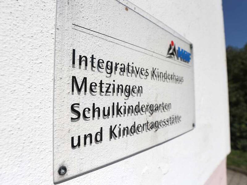 Ein Schild an einer Wand mit der Aufschrift "Integratives Kinderhaus Metzingen, Schulkindergarten und Kindertagesstätte" und einem Logo in der oberen rechten Ecke.