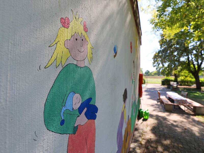 An einer Wand ist ein buntes Wandbild eines fröhlichen Kindes mit einem Baby gemalt, während im Hintergrund Tische und Bäume im Freien zu sehen sind.