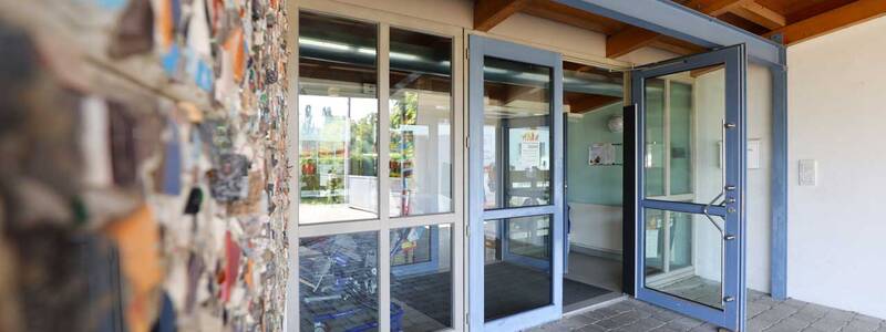 Ein Eingangsbereich mit Glas-Schwingtüren und einem Mauerstück im Vordergrund, das mit bunten Mosaikstücken verziert ist.