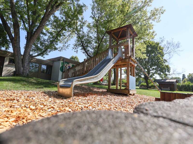 Eine hölzerne Rutsche steht auf einem Spielplatz mit hellen Holzschnitzeln umgeben von Bäumen im Hintergrund.