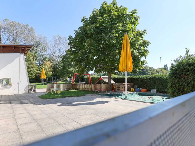 Ein Außenbereich mit einer gepflasterten Terrasse führt zu einem umzäunten Spielplatz mit zwei gelben Sonnenschirmen, einem Baum und Spielgeräten.