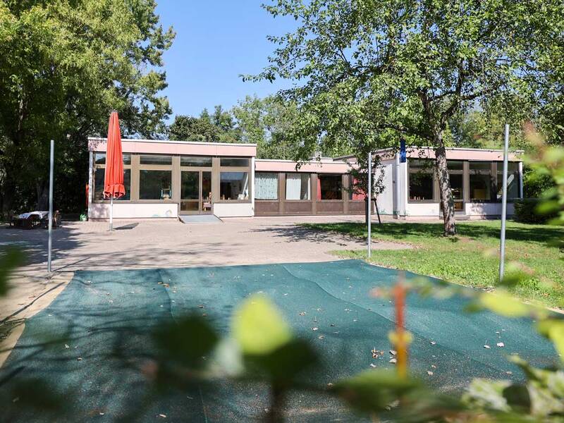 Ein modernes, einstöckiges Gebäude mit großen Glasfenstern steht inmitten einer grünen Umgebung, mit einem roten Sonnenschirm im Vordergrund.