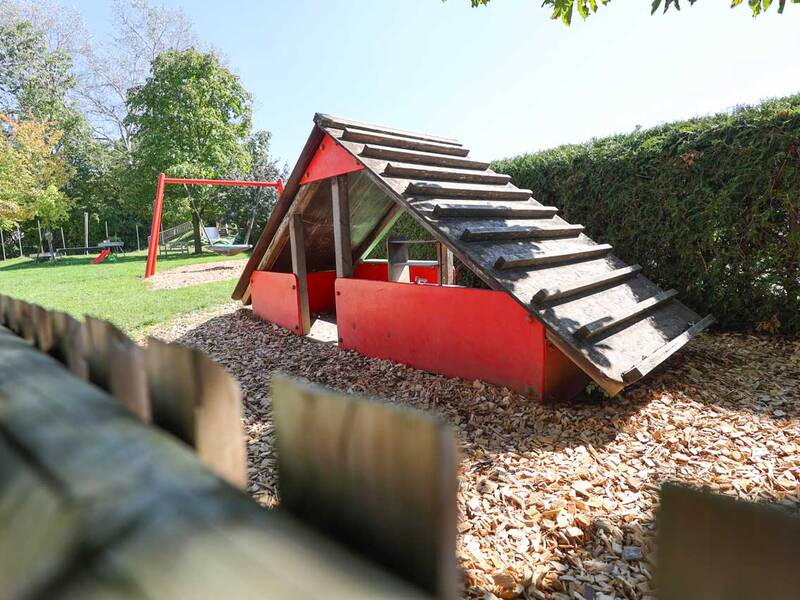 Ein Spielplatz mit einem kleinen rot-schwarzen Spielhaus aus Holz, umgeben von Rindenmulch und einer roten Schaukel im Hintergrund.