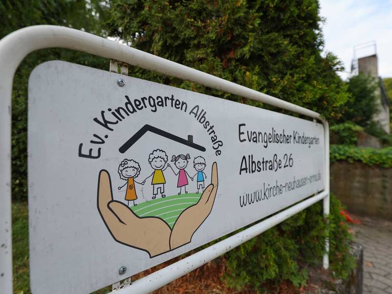 Ein Schild des Evangelischen Kindergartens Albstraße zeigt eine Zeichnung von vier lachenden Kindern, die von einem Paar Hände und einem stilisierten Hausdach umrahmt werden.