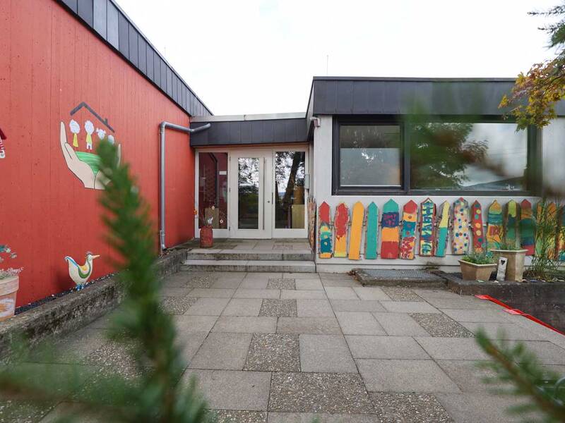 Ein einstöckiges Gebäude mit roter Fassade, bunten Zaunelementen und dekorativen Malereien steht vor einem gepflasterten Vorplatz.