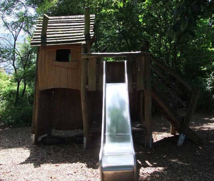 Ein hölzernes Klettergerüst mit einer Rutsche steht in einem schattigen Bereich unter Bäumen.