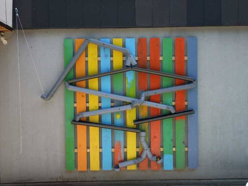 An einer Wand ist eine bunte Konstruktion aus bunten Holzlatten und grauen Rohren befestigt, die in verschiedenen Winkeln angeordnet sind.