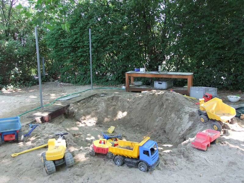 Ein Sandkasten mit Spielzeug-Trucks und Baggern, umgeben von Bäumen, mit einem Holztisch im Hintergrund, auf dem verschiedene Gegenstände stehen.