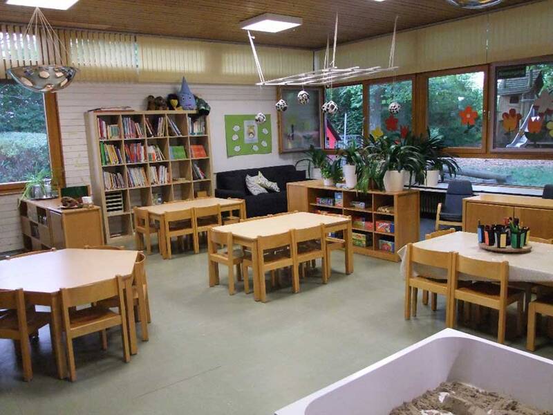 Ein heller Kindergartenraum mit mehreren kleinen Holztischen und -stühlen, Bücherregalen, Spielzeugregalen, Pflanzen und großen Fenstern mit Blick nach draußen.