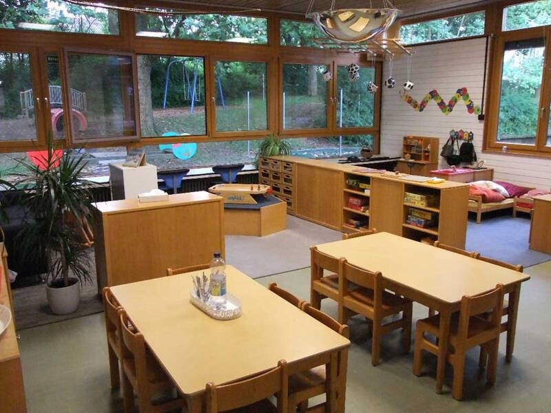 Der Raum ist ein helles, gut ausgestattetes Klassenzimmer mit hölzernen Tischen und Stühlen, Regalen voller Spiele und einem großen Fenster, das den Blick auf einen Spielplatz mit Rutsche und Schaukeln freigibt.