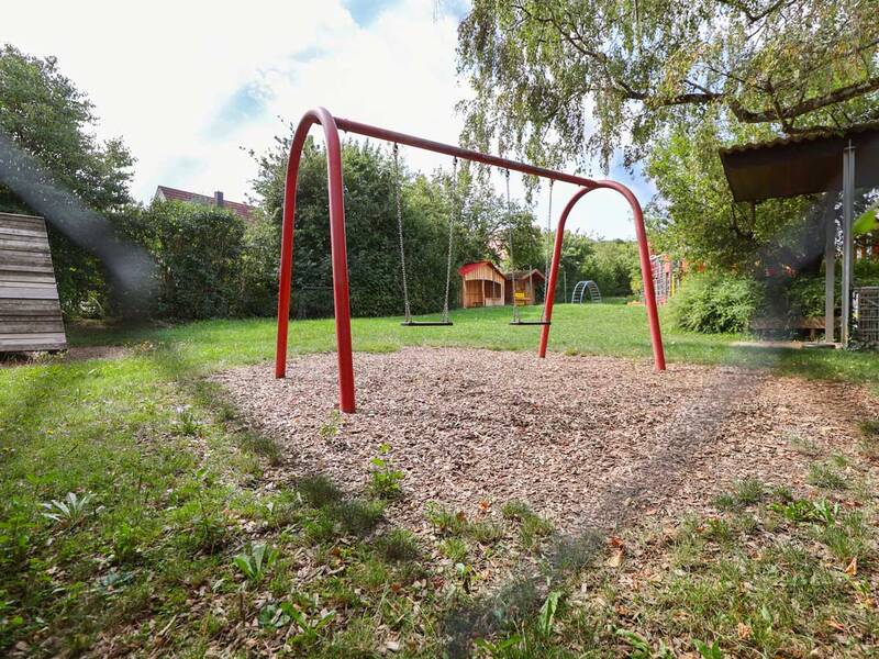 Eine rote Doppelschaukel steht auf einem Spielplatz mit Holzhäckseln, umgeben von Gras, Bäumen und einem kleinen Spielhaus im Hintergrund.